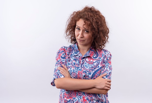 Jeune femme aux cheveux bouclés courts en chemise colorée debout avec les bras croisés sur la poitrine à la recherche avec le visage fronçant sur le mur blanc