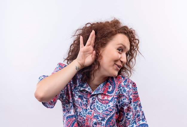 Jeune femme aux cheveux bouclés courts en chemise colorée debout avec la main près de l'oreille en essayant d'écouter quelqu'un conversation sur mur blanc