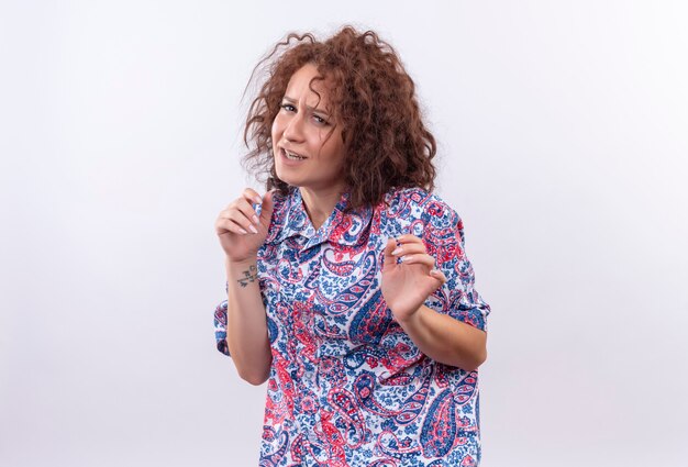 Jeune femme aux cheveux bouclés courts en chemise colorée faisant un geste de défense avec les mains avec une expression dégoûtée debout sur un mur blanc