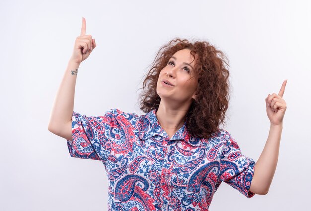 Jeune femme aux cheveux bouclés courts en chemise colorée heureux et positif regardant et pointant vers le haut avec les doigts debout sur un mur blanc