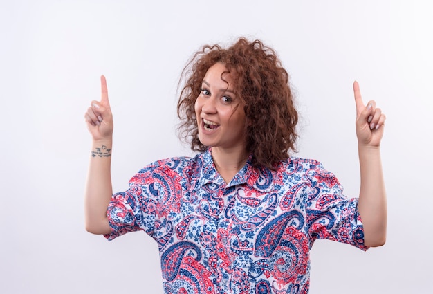 Photo gratuite jeune femme aux cheveux bouclés courts en chemise colorée heureux et positif souriant largement à la recherche et pointant vers le haut avec les doigts debout sur un mur blanc