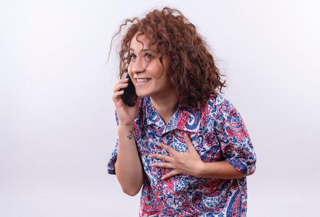 Jeune femme aux cheveux bouclés courts en chemise colorée parlant au téléphone mobile avec un sourire timide sur le visage debout sur un mur blanc