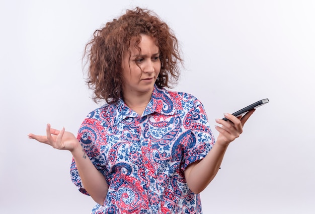 Jeune femme aux cheveux bouclés courts en chemise colorée regardant l'écran sur son téléphone mobile en écartant les bras sur les côtés à la confusion sur le mur blanc