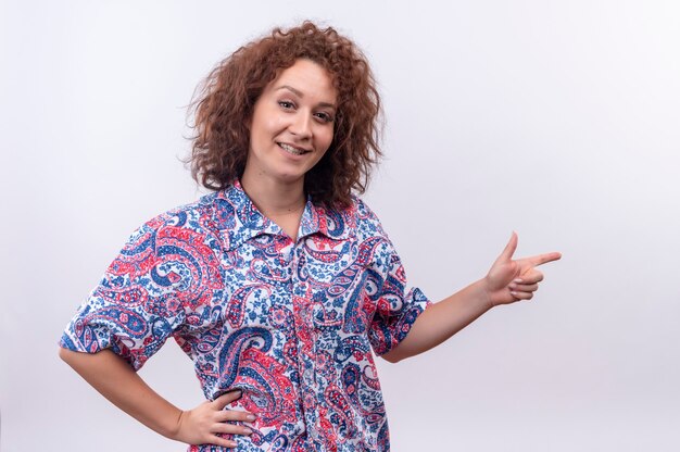 Photo gratuite jeune femme aux cheveux bouclés courts en chemise colorée smiling confiant pointant avec le doigt sur le côté sur mur blanc