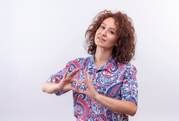 Jeune femme aux cheveux bouclés courts en chemise colorée souriant confiant montrant des signes ok avec les deux mains faisant le geste du coeur debout sur un mur blanc