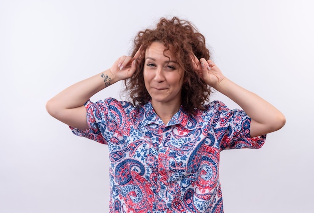 Jeune femme aux cheveux bouclés courts en chemise colorée souriant joyeusement pointant ses tempes debout sur un mur blanc