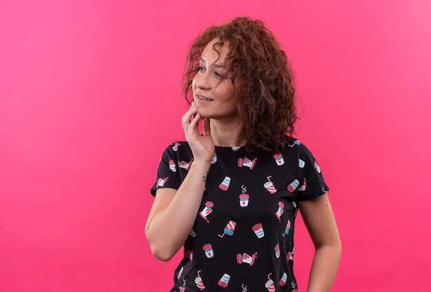 Jeune femme aux cheveux bouclés courts à côté avec une expression pensive sur le visage pensant debout sur un mur rose