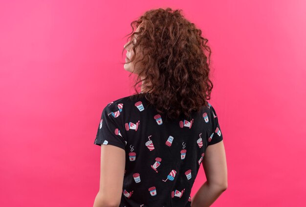 Jeune femme aux cheveux bouclés courts debout avec son dos sur un mur rose isolé
