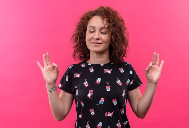 Jeune femme aux cheveux bouclés courts debout avec les yeux fermés relaxant faisant le geste de méditation avec les doigts sur le mur rose