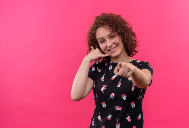 Jeune femme aux cheveux bouclés courts faisant appelez-moi geste et pointant avec le doigt à l'avant