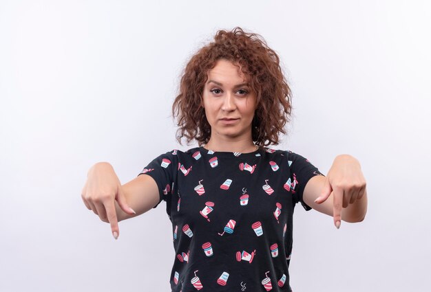 Photo gratuite jeune femme aux cheveux bouclés courts pointant les doigts vers le bas avec une expression sceptique debout sur un mur blanc