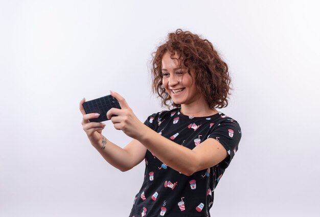 Jeune femme aux cheveux bouclés courts prenant selfie à l'aide de son smartphone souriant