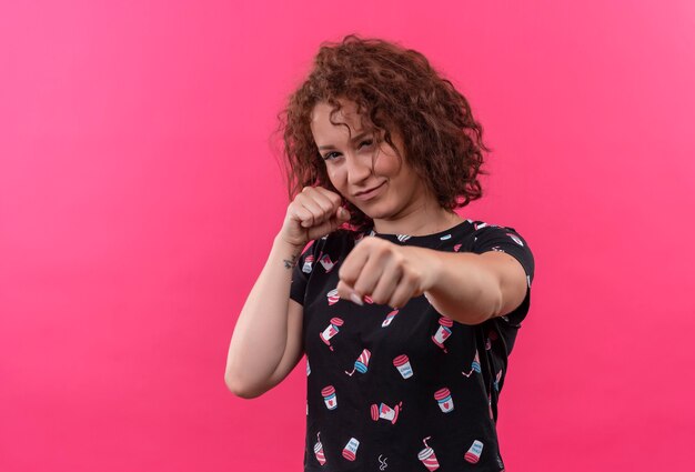 Jeune femme aux cheveux bouclés courts à la recherche de posant comme un boxeur avec les poings fermés debout sur un mur rose