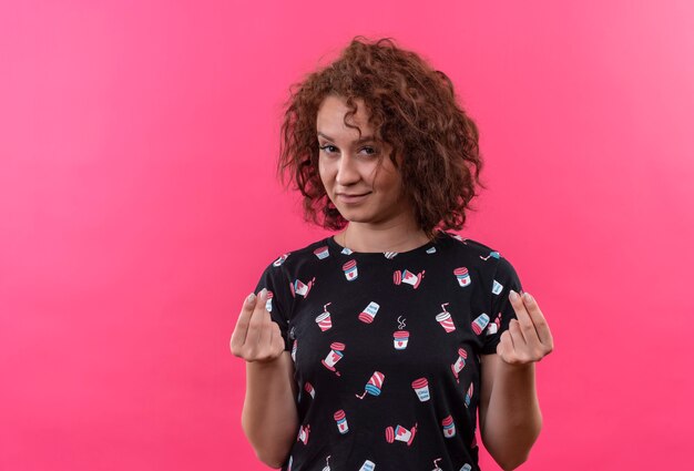 Jeune femme aux cheveux bouclés courts se frottant les doigts faisant de l'argent geste demandant de l'argent debout sur un mur rose