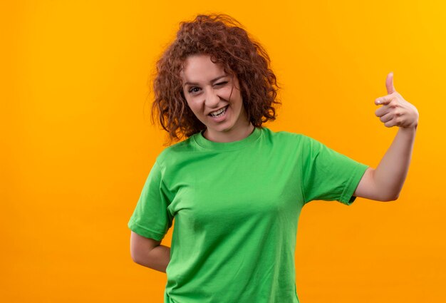 Jeune femme aux cheveux bouclés courts en t-shirt vert clignotant heureux et positif montrant les pouces vers le haut debout sur le mur orange