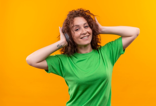 Jeune femme aux cheveux bouclés courts en t-shirt vert avec des écouteurs appréciant sa musique préférée souriant joyeusement debout sur le mur orange