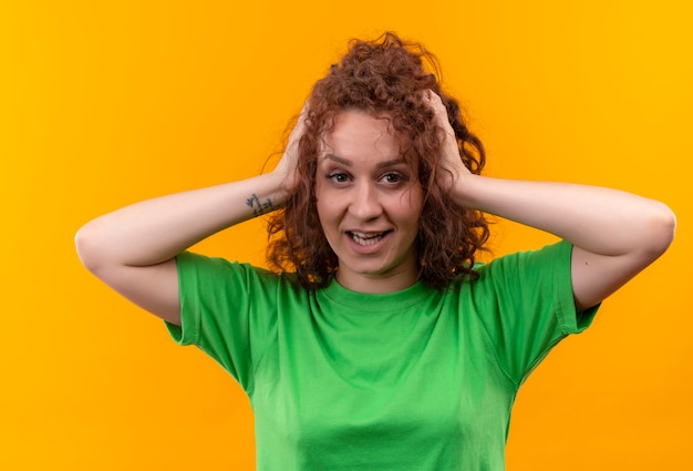 Jeune femme aux cheveux bouclés courts en t-shirt vert à la surprise de toucher ses cheveux smuling debout