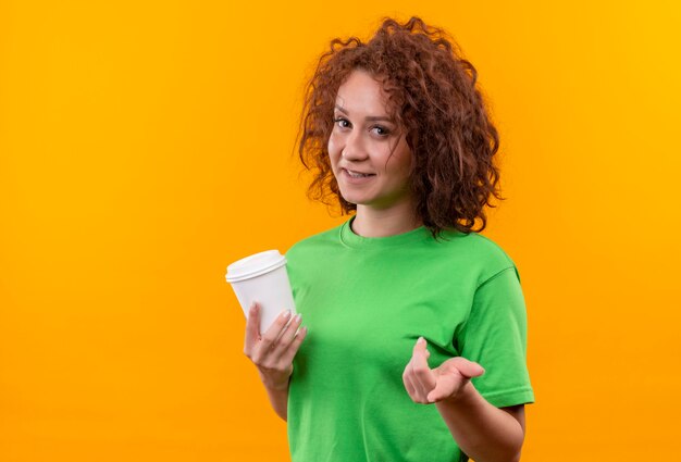 Jeune femme aux cheveux bouclés courts en t-shirt vert tenant une tasse de café en levant le bras comme posant une question debout