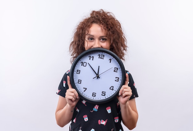 Photo gratuite jeune femme aux cheveux bouclés courts tenant horloge murale heureux souriant debout sur un mur blanc