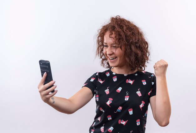 Jeune femme aux cheveux bouclés courts tenant le poing serrant le smartphone heureux et excité se réjouissant de son succès debout sur un mur blanc