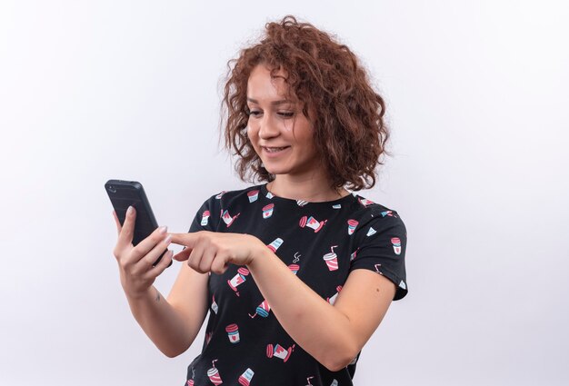 Jeune femme aux cheveux bouclés courts tenant un téléphone mobile bavardant avec quelqu'un souriant debout sur un mur blanc