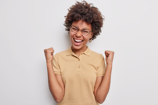 Une jeune femme aux cheveux bouclés et excitée fait un geste oui, le geste de la pompe à poing célèbre la victoire et la réussite s'exclame bruyamment des triomphes sur le succès habillé avec désinvolture isolé sur fond blanc. je suis un gagnant