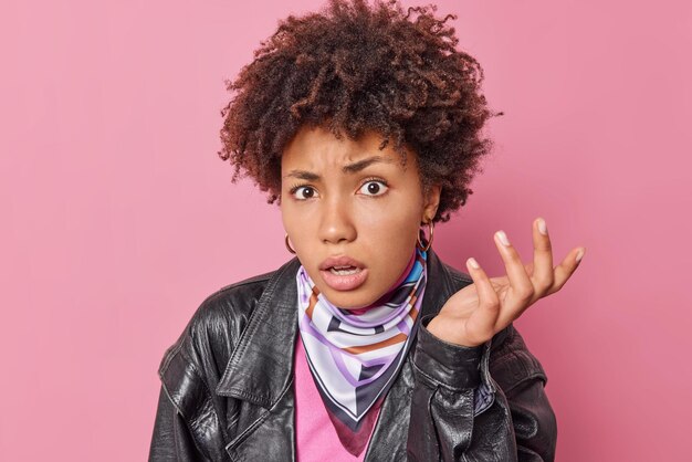 Une jeune femme aux cheveux bouclés indignée lève la paume et a l'air mécontente de hausser les épaules avec perplexité porte une veste en cuir noire isolée sur fond rose. Concept de réactions humaines. Et alors