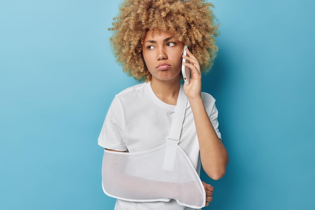 Une jeune femme aux cheveux bouclés mécontente fait un appel téléphonique au médecin a un bras cassé enveloppé dans une attelle vêtue d'un t-shirt blanc décontracté regarde ailleurs pose sur fond bleu Problèmes de santé