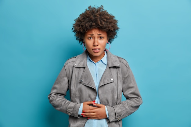 Photo gratuite une jeune femme aux cheveux bouclés mécontente ressent une gêne dans le ventre, a la diarrhée, fronce les sourcils, presse les mains sur le ventre, a des problèmes de santé et des troubles de l'alimentation