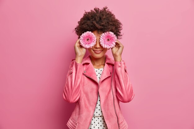 jeune femme aux cheveux bouclés tient une fleur de gerbera rose, couvre les yeux, vêtue d'une veste rose à la mode, fait de la décoration, pose à l'intérieur.