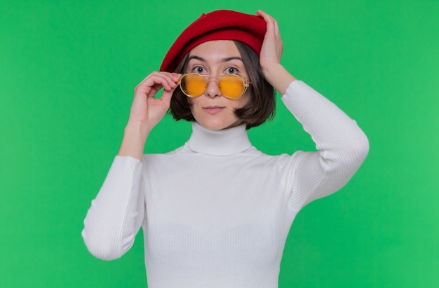 Jeune femme aux cheveux courts en col roulé blanc portant un béret et des lunettes de soleil jaunes à l'avant debout sur un mur confus vert