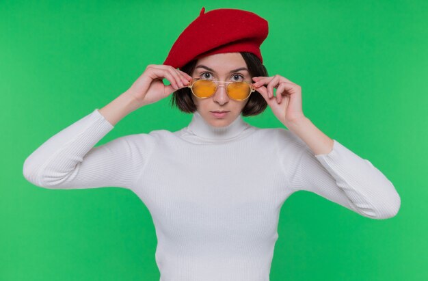 Jeune femme aux cheveux courts en col roulé blanc portant béret et lunettes de soleil jaunes