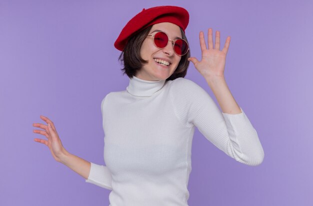 Jeune femme aux cheveux courts en col roulé blanc portant béret et lunettes de soleil rouges heureux et excité souriant gaiement gesticulant avec les mains debout sur le mur bleu