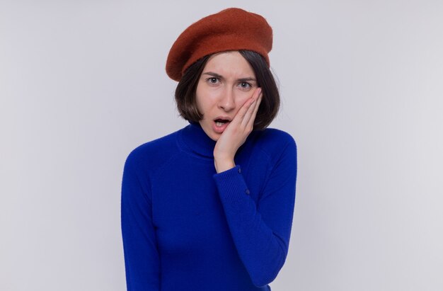 Jeune femme aux cheveux courts en col roulé bleu portant un béret à l'avant d'être choqué couvrant la bouche avec la main debout sur un mur blanc