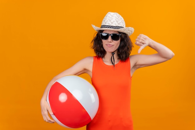 Une jeune femme aux cheveux courts dans une chemise orange portant un chapeau et des lunettes de soleil tenant ballon gonflable montrant les pouces vers le bas