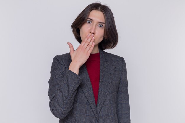 Jeune femme aux cheveux courts portant une veste grise à l'avant d'être choqué couvrant la bouche avec la main debout sur un mur blanc