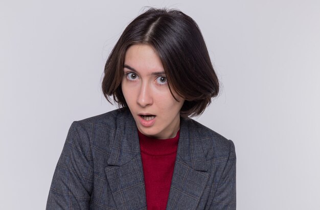 Jeune femme aux cheveux courts portant une veste grise à l'avant surpris et intrigué debout sur un mur blanc