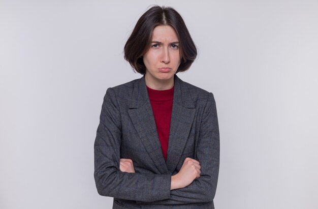 Jeune femme aux cheveux courts portant une veste grise à l'avant avec le visage fronçant étant offensé avec les bras croisés debout sur un mur blanc