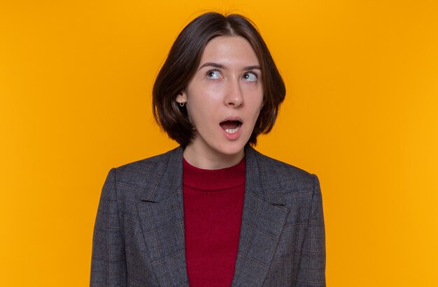 Jeune femme aux cheveux courts portant une veste grise à la surprise debout sur un mur orange