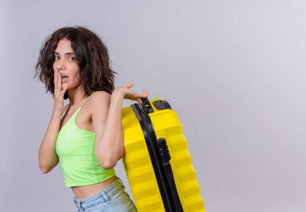 Une jeune femme aux cheveux courts en vert crop top en gardant la main sur la bouche et tenant une valise jaune sur fond blanc