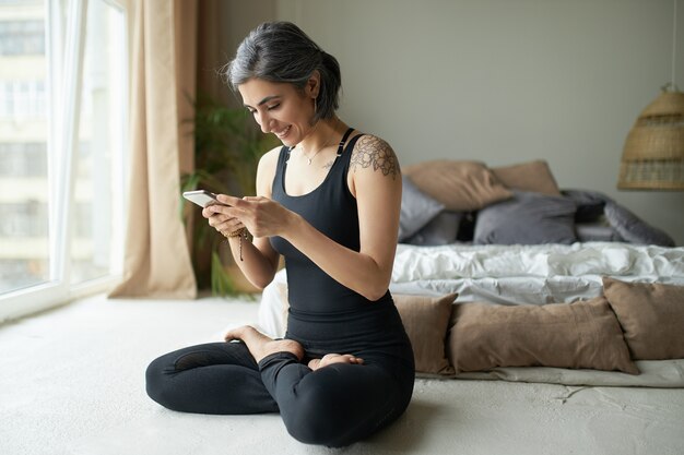 Jeune femme aux cheveux grisâtres et tatouage pratiquant le yoga et utilisant le smartphone