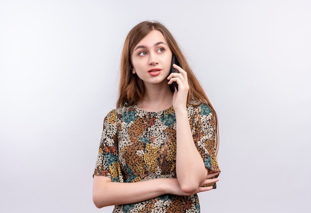 Jeune femme aux cheveux longs portant une robe colorée souriant tout en parlant au téléphone mobile debout sur un mur blanc