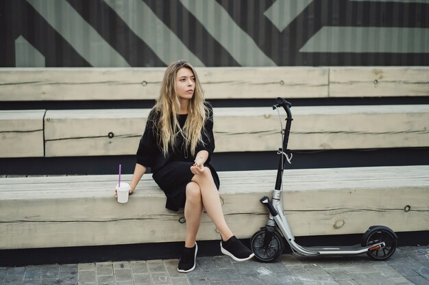 Jeune femme aux cheveux longs sur un scooter électrique. La fille sur le scooter électrique boit du café.