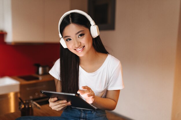 Jeune femme aux cheveux noirs avec le sourire regarde en avant, tient la tablette et écoute de la musique avec des écouteurs