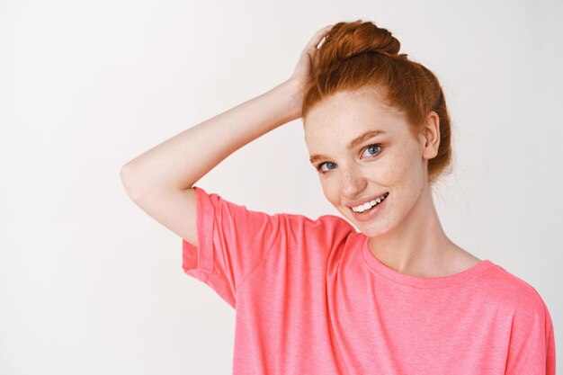 Jeune femme aux cheveux rouges peignés en chignon désordonné souriante mignonne à l'avant, montrant une peau éclatante de santé sans acné ni imperfections, mur blanc