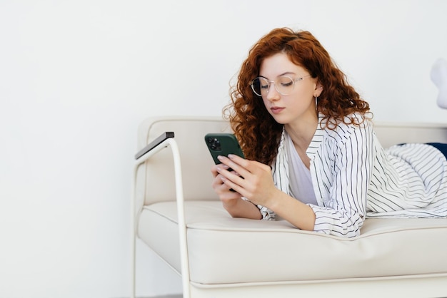 Jeune femme aux cheveux rouges utilisant un téléphone intelligent tout en se reposant sur un canapé à la maison