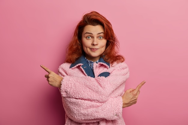 Jeune femme aux cheveux roux a une expression de visage hésitante surprenante, pointe de différents côtés, fait le choix entre deux options, porte un manteau rose