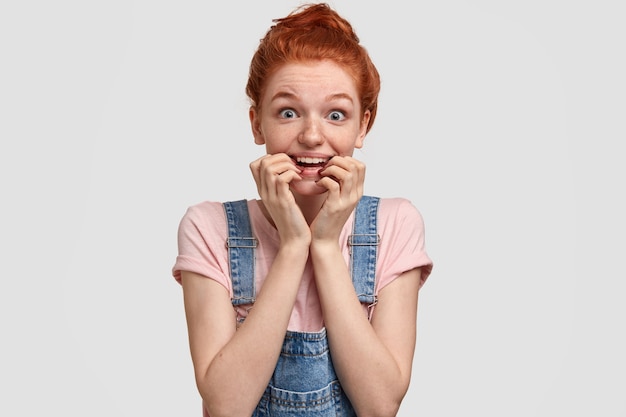 Photo gratuite jeune femme aux cheveux roux perplexe avec une expression faciale surprise inquiète, garde les mains près de la bouche, regarde avec choc, a un nœud de cheveux foxy, pose contre un mur blanc. concept de réaction