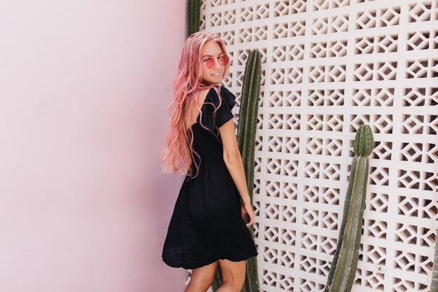 jeune femme aux longs cheveux roses ondulés posant avec sourire à côté de cactus.