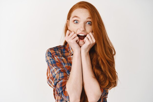 Jeune femme aux longs cheveux roux et aux yeux bleus regardant avec admiration et étonnement la caméra appuyée sur les mains et regardant fascinée debout sur fond blanc